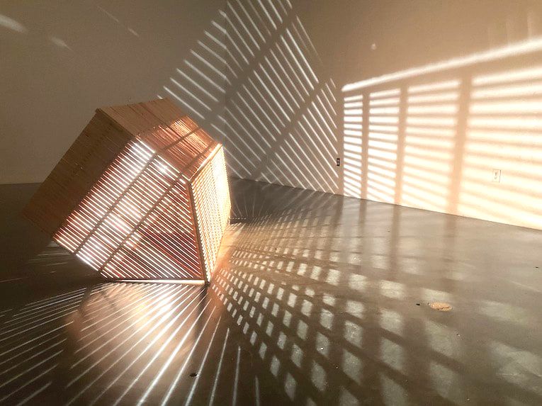 Wooden sculpture of open slatted barn with light inside radiating patterns into a darkened room. 